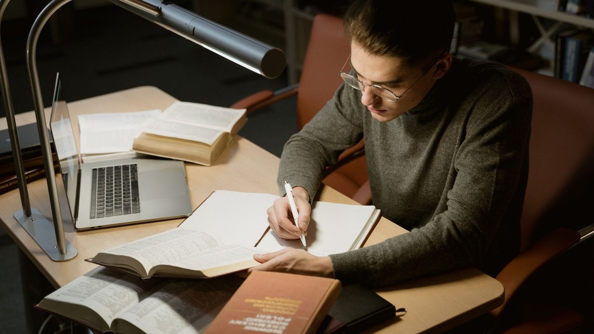 Persona tomando notas mientras contrasta un libro y un portátil