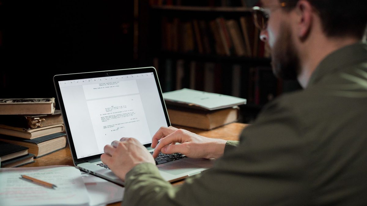 Persona escribiendo en un portátil rodeada de libros y documentos apilados